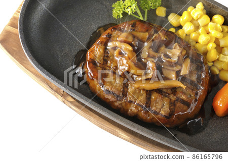 A bird's-eye view of a hot Japanese-style hamburger steak with parsley, carrot glass, corn, and mushroom sauce on an iron plate on a white background. A bird's-eye view of a hot Japanese-style hamburger steak with parsley, carrot glass, corn, and mushroom sauce on an iron plate on a white background. 86165796