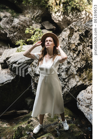 Beautiful woman in white long dress and hat is standing in forest on the stone. Travel concept. Beautiful woman in white long dress and hat is standing in forest on the stone. Travel concept. 86169653
