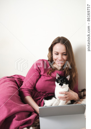 Young millennial woman in pink dress with long hair using a tablet and watcing movies with her black and white tuxedo cat at home. Young millennial woman in pink dress with long hair using a tablet and watcing movies with her black and white tuxedo cat at home. 86169757