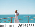 Young woman in white dress and hat stands by the sea. 86169783