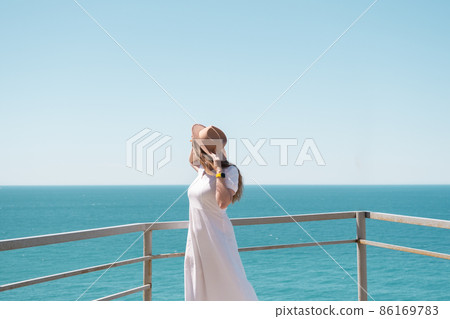 Young woman in white dress and hat stands by the sea. 86169783