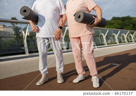 Mature couple in sportswear with mats ready for workout on footbridge at sunset closeup Mature couple in sportswear with mats ready for workout on footbridge at sunset closeup 86170137