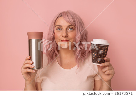 Portrait of positive caucasian woman comparing reusable thermo cup with disposable paper coffee cup. Zero waste and eco friendly concept Portrait of positive caucasian woman comparing reusable thermo cup with disposable paper coffee cup. Zero waste and eco friendly concept 86173571