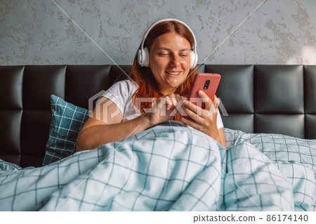 Woman in pajamas wearing headphones listening to music on smartphone relaxing sitting on bed at home bedroom 86174140
