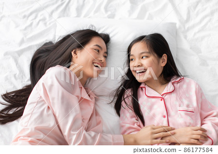 Happy pretty korean millennial lady and teen girl in pink pajamas lie on white bed in bedroom, top view Happy pretty korean millennial lady and teen girl in pink pajamas lie on white bed in bedroom, top view 86177584
