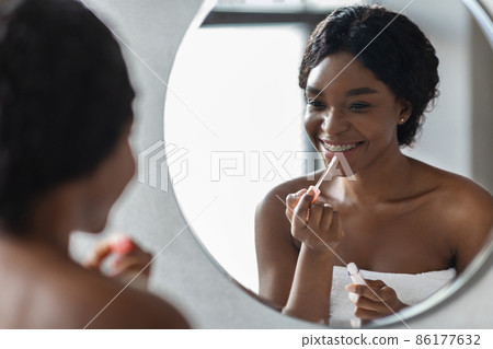 Smiling black woman applying lip gloss, looking at mirror 86177632