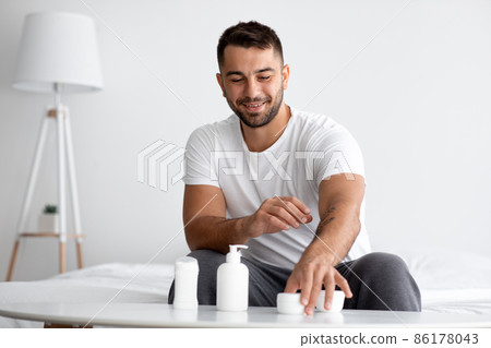Smiling young caucasian muscular handsome man at table with jars of different moisturizing anti-aging cosmetics Smiling young caucasian muscular handsome man at table with jars of different moisturizing anti-aging cosmetics 86178043