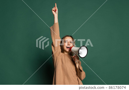 Portrait of young beautiful girl wearing brown sweatshirt shouting at megaphone isolated on green background. Concept of emotions, 86178074