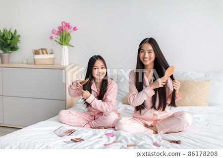 Happy japanese teenage girl and millennial female brushing long smooth hair on bed with cosmetics 86178208
