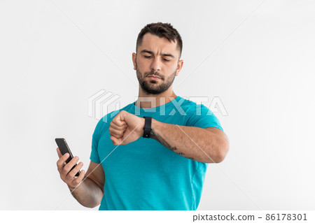Serious young caucasian man looking at fitness tracker and hold smartphone, check pulse and sport result 86178301