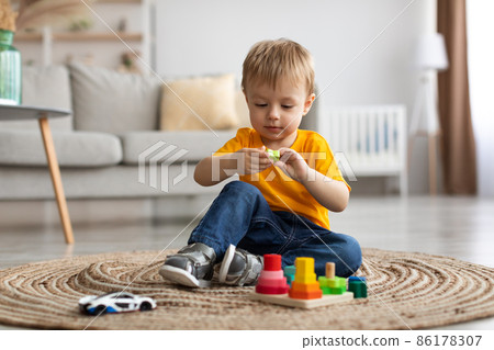 Child development games. Engaged toddler boy playing with wooden colorful stacking and sorting toy, sitting on carpet Child development games. Engaged toddler boy playing with wooden colorful stacking and sorting toy, sitting on carpet 86178307