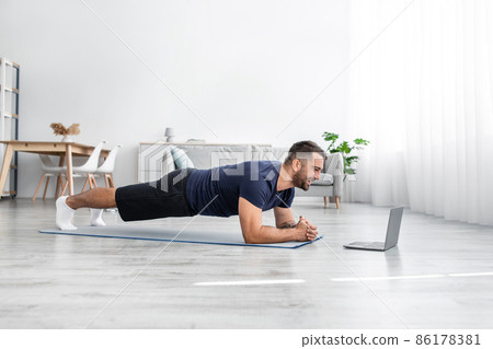 Smiling young caucasian guy doing plank on mat on floor and look at laptop in living room interior, profile Smiling young caucasian guy doing plank on mat on floor and look at laptop in living room interior, profile 86178381