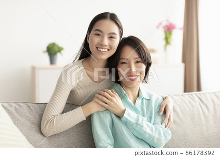 Happy mature Asian woman and her adult daughter hugging and smiling at camera, sitting on sofa at home Happy mature Asian woman and her adult daughter hugging and smiling at camera, sitting on sofa at home 86178392