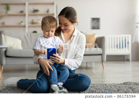 Young mother and her child boy sitting on floor carpet with smartphone, watching video or cartoons from internet Young mother and her child boy sitting on floor carpet with smartphone, watching video or cartoons from internet 86178394