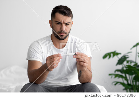 Sad young european muscular male sits on white bed, looks at thermometer with high temperature in bedroom 86178396