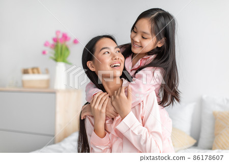 Glad smiling pretty cute asian teenage girl hugging millennial woman in pink pajamas in bedroom interior 86178607