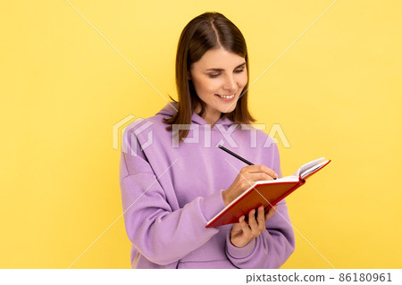 Portrait of happy inspired woman smiling and writing down interesting idea in notebook, making plans, to-do list, wearing purple hoodie. Indoor studio shot isolated on yellow background. Portrait of happy inspired woman smiling and writing down interesting idea in notebook, making plans, to-do list, wearing purple hoodie. Indoor studio shot isolated on yellow background. 86180961
