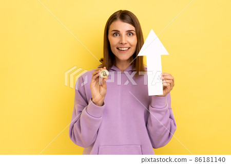 Portrait of young adult attractive dark haired woman showing growth increase of bitcoin, expressing positive emotions, wearing purple hoodie. Indoor studio shot isolated on yellow background. 86181140