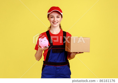 Portrait of smiling positive courier woman standing holding cardboard box and piggybank, looking at camera with happy expression, wearing overalls. Indoor studio shot isolated on yellow background. 86181176