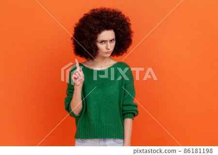 Portrait of serious woman with Afro hairstyle wearing green casual style sweater looking at camera, showing warning attention sign. Indoor studio shot isolated on orange background. Portrait of serious woman with Afro hairstyle wearing green casual style sweater looking at camera, showing warning attention sign. Indoor studio shot isolated on orange background. 86181198