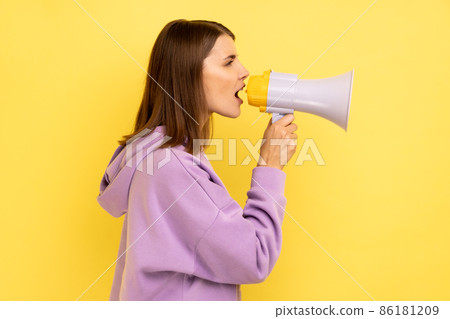 Side view portrait of young adult beautiful woman screaming in megaphone and announcing discounts in mall, wearing purple hoodie. Indoor studio shot isolated on yellow background. Side view portrait of young adult beautiful woman screaming in megaphone and announcing discounts in mall, wearing purple hoodie. Indoor studio shot isolated on yellow background. 86181209