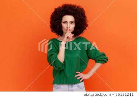 Portrait of serious woman with Afro hairstyle wearing green casual style sweater standing with finger near lips, showing silent gesture. Indoor studio shot isolated on orange background. Portrait of serious woman with Afro hairstyle wearing green casual style sweater standing with finger near lips, showing silent gesture. Indoor studio shot isolated on orange background. 86181211