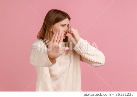 Portrait of woman pinching her nose, holding breath to avoid bad smell and showing stop gesture, disgusted by stinky fart odor, wearing white sweater. Indoor studio shot isolated on pink background. Portrait of woman pinching her nose, holding breath to avoid bad smell and showing stop gesture, disgusted by stinky fart odor, wearing white sweater. Indoor studio shot isolated on pink background. 86181223