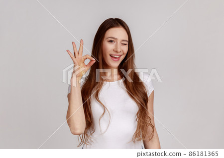 Portrait of woman gesturing okay sign, completely agree with suggestion, confident in success, expressing playful emotions, wearing white T-shirt. Indoor studio shot isolated on gray background. Portrait of woman gesturing okay sign, completely agree with suggestion, confident in success, expressing playful emotions, wearing white T-shirt. Indoor studio shot isolated on gray background. 86181365