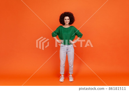 Full length portrait of positive woman with Afro hairstyle wearing green casual style sweater standing with hands on hips and toothy smile. Indoor studio shot isolated on orange background. Full length portrait of positive woman with Afro hairstyle wearing green casual style sweater standing with hands on hips and toothy smile. Indoor studio shot isolated on orange background. 86181511
