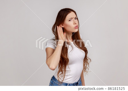 Portrait of curious woman holding hand near ear trying to hear secrets and find out gossips, listen to private whispers, wearing white T-shirt. Indoor studio shot isolated on gray background. Portrait of curious woman holding hand near ear trying to hear secrets and find out gossips, listen to private whispers, wearing white T-shirt. Indoor studio shot isolated on gray background. 86181525