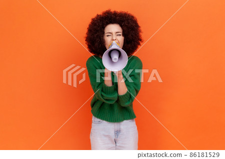 Attractive woman with Afro hairstyle wearing green casual style sweater screaming loud using megaphone, making announcement. presentation. Indoor studio shot isolated on orange background. 86181529