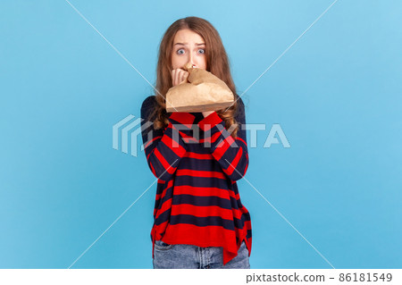 Sick woman wearing striped casual style sweater, breathing into paper bag to improve well-being, overcoming stress, looking at camera with fear. Indoor studio shot isolated on blue background. Sick woman wearing striped casual style sweater, breathing into paper bag to improve well-being, overcoming stress, looking at camera with fear. Indoor studio shot isolated on blue background. 86181549