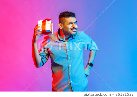 Portrait of funny man in shirt holding present box in hand, being interested what inside, frowning face, has insidious expression. Indoor studio shot isolated on colorful neon light background. Portrait of funny man in shirt holding present box in hand, being interested what inside, frowning face, has insidious expression. Indoor studio shot isolated on colorful neon light background. 86181552