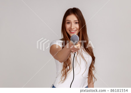 Portrait of smiling attractive woman with dark hair standing offering microphone, journalist taking interview, wearing white T-shirt. Indoor studio shot isolated on gray background. Portrait of smiling attractive woman with dark hair standing offering microphone, journalist taking interview, wearing white T-shirt. Indoor studio shot isolated on gray background. 86181554