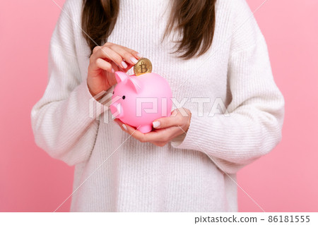 Close up of unknown faceless woman looking at camera, putting coin into piggybank, savings of money, wearing white casual style sweater. Indoor studio shot isolated on pink background. 86181555
