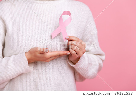 Closeup of unknown female holding pink ribbon, symbol of breast cancer awareness, cancer prevention, wearing white casual style sweater. Indoor studio shot isolated on pink background. 86181558