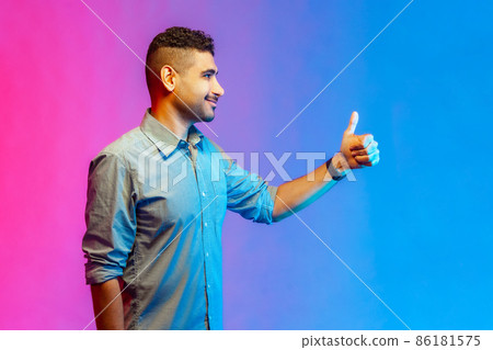 Side view portrait of smiling man in shirt showing thumb up, like gesture, looking ahead, demonstrating his satisfaction. Indoor studio shot isolated on colorful neon light background. Side view portrait of smiling man in shirt showing thumb up, like gesture, looking ahead, demonstrating his satisfaction. Indoor studio shot isolated on colorful neon light background. 86181575