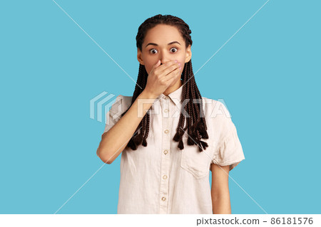 Worried beautiful woman with dreadlocks wants to scream, covers mouth with palm, stares at something terrible, has eyes popped out, wearing white shirt. Indoor studio shot isolated on blue background. Worried beautiful woman with dreadlocks wants to scream, covers mouth with palm, stares at something terrible, has eyes popped out, wearing white shirt. Indoor studio shot isolated on blue background. 86181576