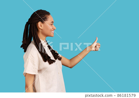 Side view portrait of woman with black dreadlocks smiling happily, keeps thumbs up, approves something, has cheerful expression, wearing white shirt. Indoor studio shot isolated on blue background. Side view portrait of woman with black dreadlocks smiling happily, keeps thumbs up, approves something, has cheerful expression, wearing white shirt. Indoor studio shot isolated on blue background. 86181579