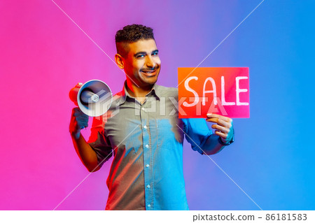 Portrait of happy delighted man in shirt holding sale card and megaphone, announcing discounts in shopping mall, looking at camera. Indoor studio shot isolated on colorful neon light background. 86181583