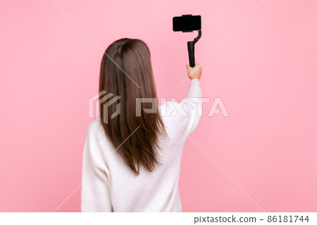 Back view of female broadcasting livestream, using steadicam and phone, posing backwards to camera, wearing white casual style sweater. Indoor studio shot isolated on pink background. 86181744