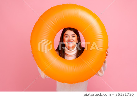 Young adult woman with happy expression looking trough orange rubber ring, enjoying her vacation, wearing white casual style sweater. Indoor studio shot isolated on pink background. Young adult woman with happy expression looking trough orange rubber ring, enjoying her vacation, wearing white casual style sweater. Indoor studio shot isolated on pink background. 86181750