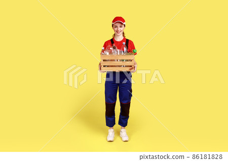 Full length portrait of positive satisfied woman volunteer standing with box full of empty plastic bottles, wearing overalls and red cap. Indoor studio shot isolated on yellow background. Full length portrait of positive satisfied woman volunteer standing with box full of empty plastic bottles, wearing overalls and red cap. Indoor studio shot isolated on yellow background. 86181828