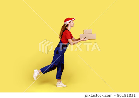 Side view of courier woman being hurry to deliver two cardboard parcels, fast delivery door-to-door, wearing overalls and red cap. Indoor studio shot isolated on yellow background. Side view of courier woman being hurry to deliver two cardboard parcels, fast delivery door-to-door, wearing overalls and red cap. Indoor studio shot isolated on yellow background. 86181831