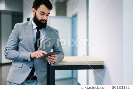 Confident adult businessman surfing tablet while leaning on table 86183321
