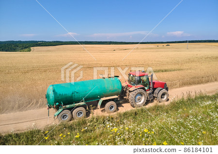 Tractor with water tank trailer driving on dirt road between agricultural fields during harvest season Tractor with water tank trailer driving on dirt road between agricultural fields during harvest season 86184101