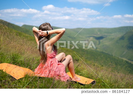 Young woman in red dress resting on green grassy field on a sunny day in summer mountains enjoying view of nature. 86184127