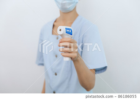 Temperature measurement gun in doctor hands. Close-up shot of doctor wearing protective surgical mask ready to use infrared isometric thermometer gun to check body temperature for virus symptoms. Temperature measurement gun in doctor hands. Close-up shot of doctor wearing protective surgical mask ready to use infrared isometric thermometer gun to check body temperature for virus symptoms. 86186055