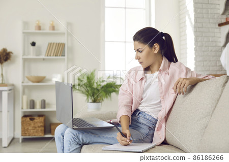 Woman taking online course. Serious focused young lady sitting on couch at home, watching webinar on laptop computer, using modern digital learning tools, doing research and taking study notes Woman taking online course. Serious focused young lady sitting on couch at home, watching webinar on laptop computer, using modern digital learning tools, doing research and taking study notes 86186266