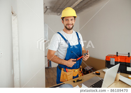 Professional handyman worker in uniform wearing tool belt and hardhat smiling at camera, holding hammer during renovation work indoors Professional handyman worker in uniform wearing tool belt and hardhat smiling at camera, holding hammer during renovation work indoors 86189470
